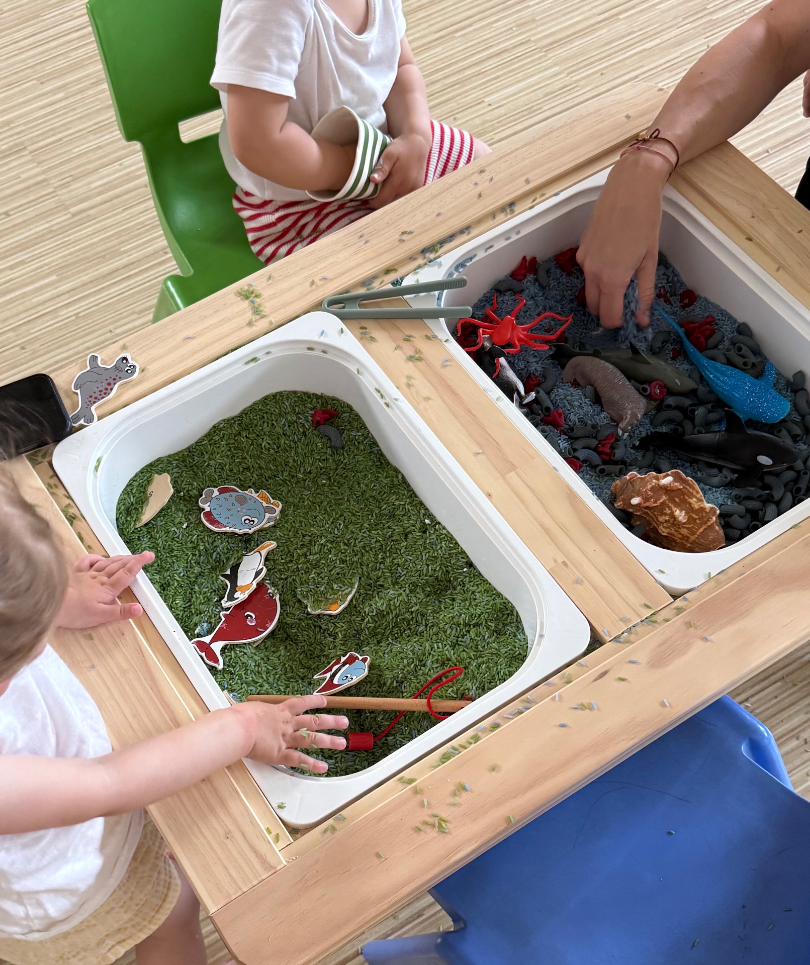 Děti a dospělý u dřevěného stolu objevují dvě smyslové vaničky – jednu se zelenou rýží a druhou s šedým materiálem a hračkami